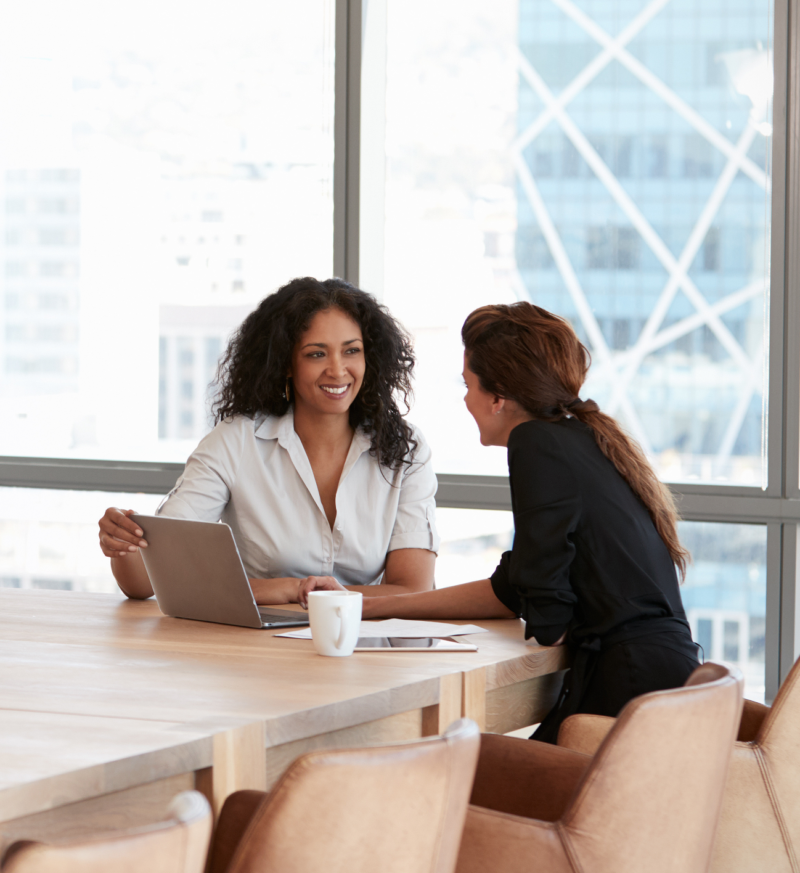 Two women smiling and talking at a table with a laptop in front of them