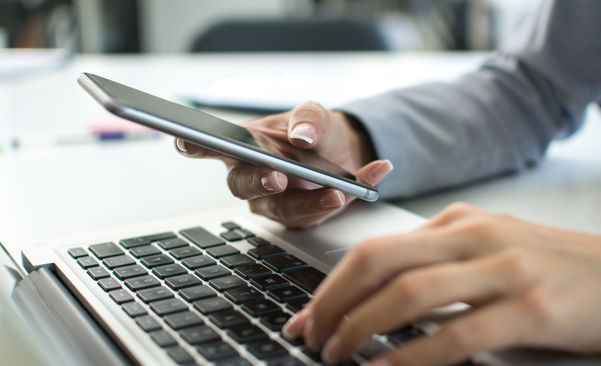 A person's hand holding a mobile phone while typing on a laptop