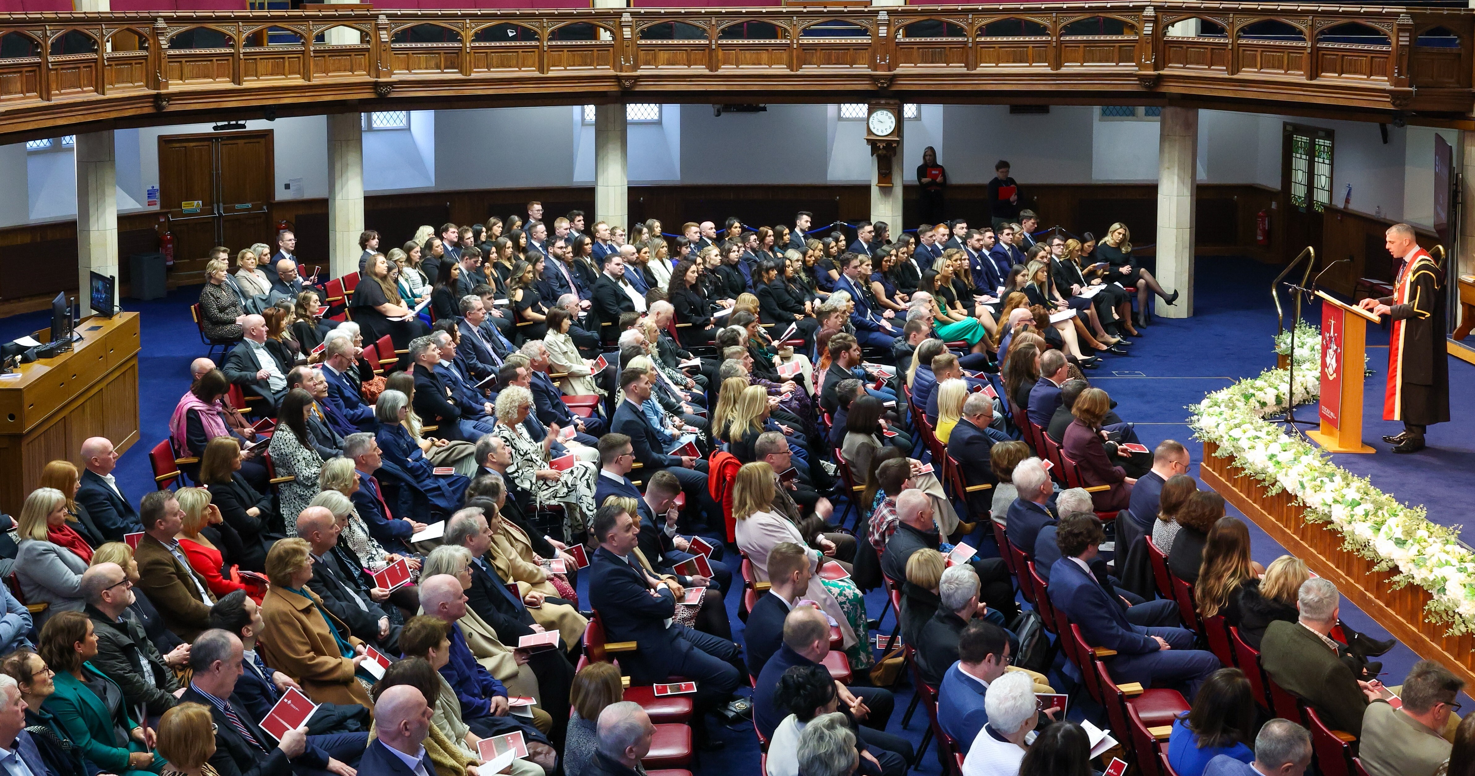 An audience sitting in a large ceremony room as a speaker speaks at a podium on the stage