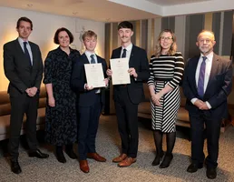 Two competition winners holding certificates flanked by competition judges