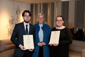 3 people standing indoors, 2 holding certificates