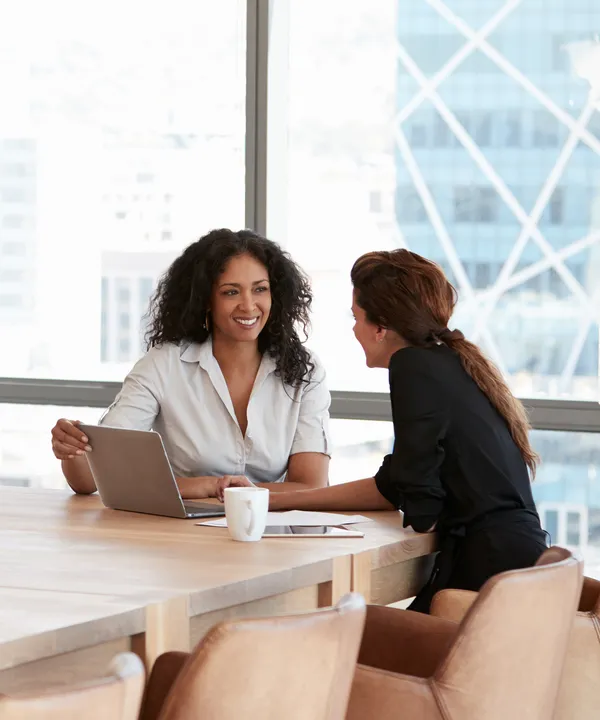 Two women smiling and talking at a table with a laptop in front of them